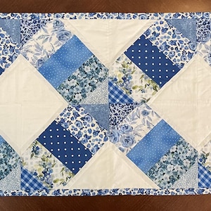 May include: A rectangular table runner with a patchwork design. The runner features a pattern of alternating white squares and blue patterned rectangles. The blue patterns include floral, polka dot, and gingham designs. The edges are finished with a patterned border.