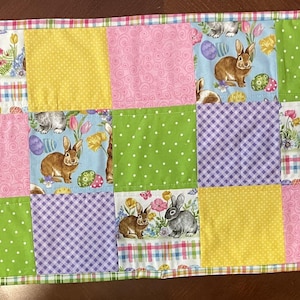 May include: A colorful Easter-themed table runner. The patchwork design features squares of green polka dots, purple gingham, yellow, and pink fabric. Some squares have bunny and floral prints. The edges are trimmed with a plaid pattern.