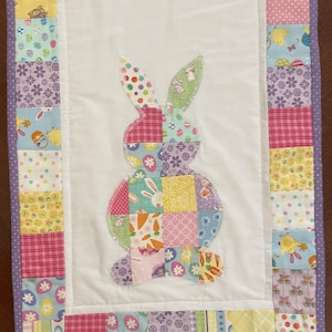 May include: A quilted Easter table runner featuring a patchwork bunny applique. The bunny is made of various patterned fabrics in pastel colors. The border is a mix of patterned squares in pink, yellow, and blue, with a purple polka dot trim.
