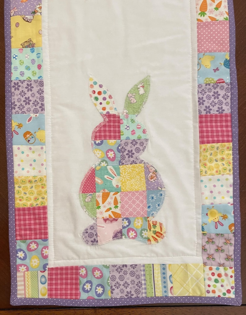 May include: A quilted Easter table runner featuring a patchwork bunny applique. The bunny is made of various patterned fabrics in pastel colors. The border is a mix of patterned squares in pink, yellow, and blue, with a purple polka dot trim.