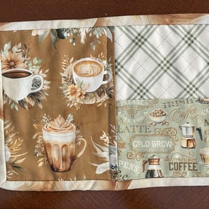 May include: A quilted coffee-themed placemat featuring three coffee cup illustrations on a brown floral background. The design includes a plaid pattern and a section with coffee-related text and images. The edges are finished with a cream-colored border.