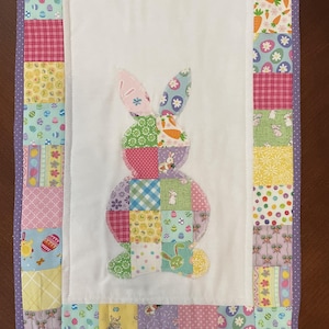 May include: A colorful Easter-themed table runner featuring a patchwork design. The center showcases a bunny applique made of various patterned fabrics. The border is adorned with coordinating fabrics in pastel colors, including pink, yellow, and purple. The runner is approximately 12 inches wide and 36 inches long.
