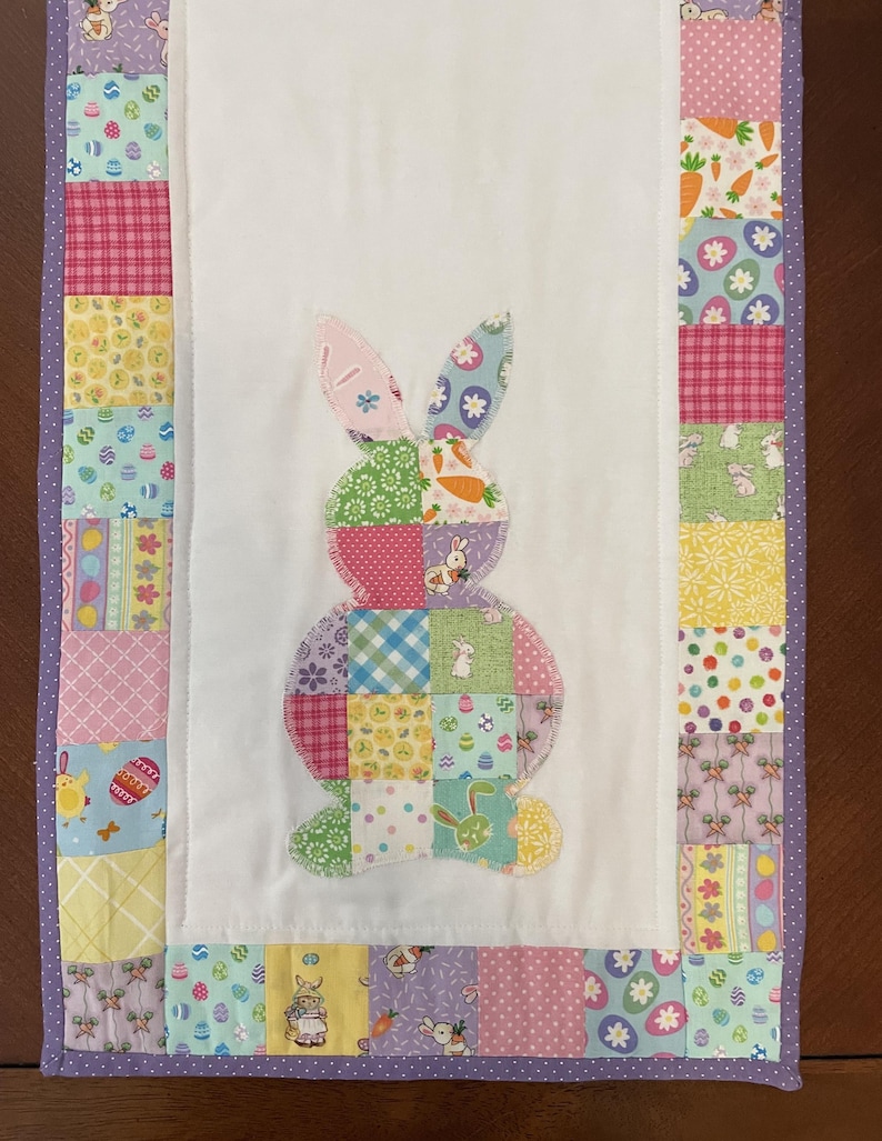 May include: A colorful Easter-themed table runner featuring a patchwork design. The center showcases a bunny applique made of various patterned fabrics. The border is adorned with coordinating fabrics in pastel colors, including pink, yellow, and purple. The runner is approximately 12 inches wide and 36 inches long.