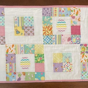 May include: A rectangular Easter-themed quilt with a white background and colorful patchwork squares. The quilt features egg appliqués and a pink polka-dot border. The patchwork includes bunny and carrot patterns.