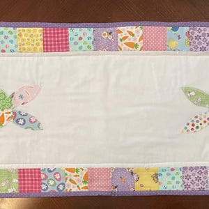 May include: A rectangular Easter-themed table runner with a white center and a patchwork border. The border features squares of various pastel fabrics. Two bunny shapes are appliqued on the white center, made of colorful patchwork. The runner is trimmed with a purple border.