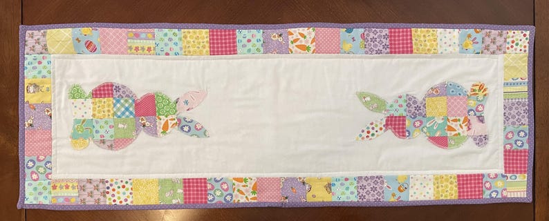 May include: A rectangular Easter-themed table runner with a white center and a patchwork border. The border features squares of various pastel fabrics. Two bunny shapes are appliqued on the white center, made of colorful patchwork. The runner is trimmed with a purple border.