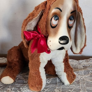 May include: A vintage plush dog toy with a painted face, brown fur, and a red bow. The dog has large, droopy ears and a white chest and paws. The toy is sitting on a lace tablecloth.