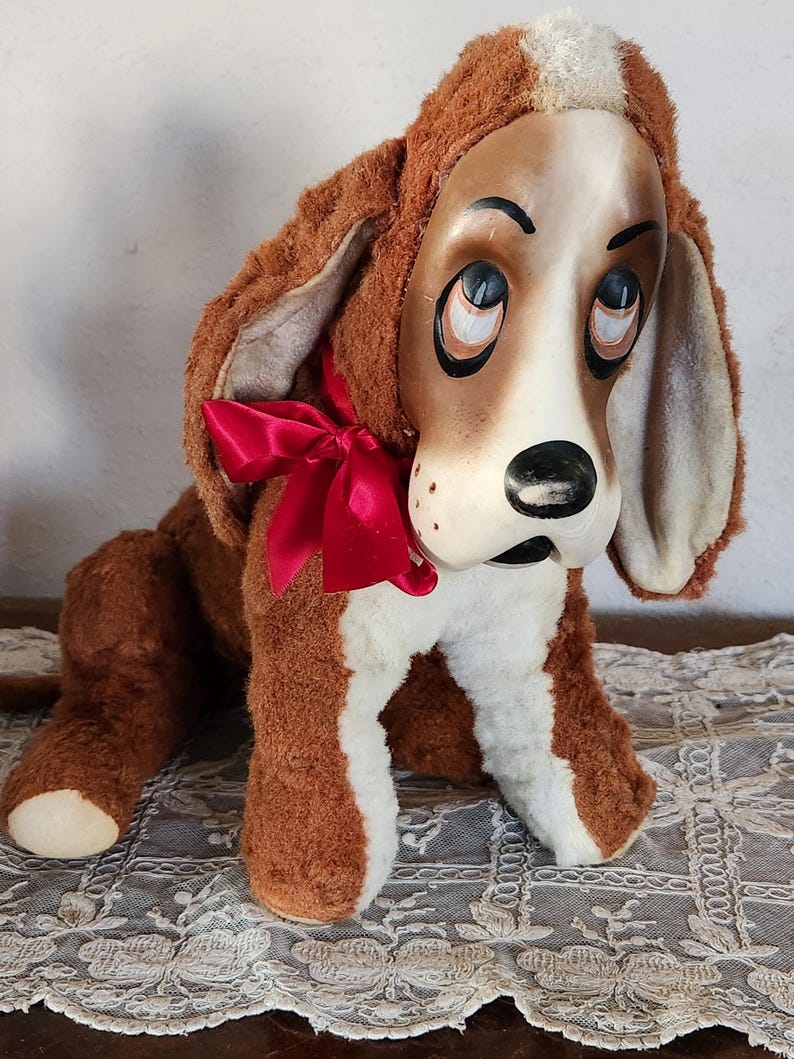 May include: A vintage plush dog toy with a painted face, brown fur, and a red bow. The dog has large, droopy ears and a white chest and paws. The toy is sitting on a lace tablecloth.