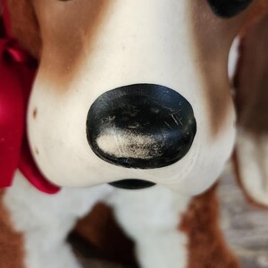 May include: Close-up of a vintage dog figurine. The dog has brown and white fur, with a black nose and large, dark eyes. A red ribbon is tied around its neck. The figurine is likely made of ceramic or a similar material.