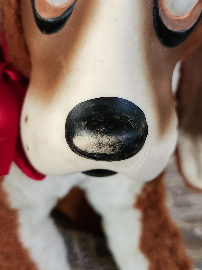 May include: Close-up of a vintage dog figurine. The dog has brown and white fur, with a black nose and large, dark eyes. A red ribbon is tied around its neck. The figurine is likely made of ceramic or a similar material.