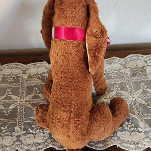 May include: A vintage brown plush basset hound dog toy with long floppy ears and a red ribbon around its torso. The dog has a white patch on its head and is sitting on a white lace tablecloth.