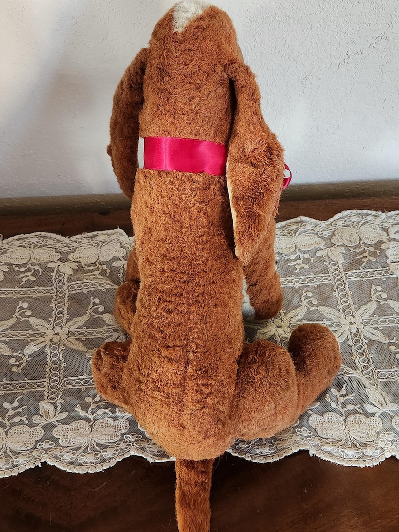 May include: A vintage brown plush basset hound dog toy with long floppy ears and a red ribbon around its torso. The dog has a white patch on its head and is sitting on a white lace tablecloth.
