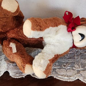 May include: A vintage brown and white plush toy bear with a red bow. The bear has a cream-colored face and paws. The bear is lying on a lace-trimmed surface.