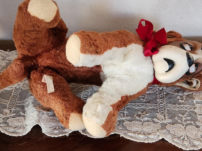 May include: A vintage brown and white plush toy bear with a red bow. The bear has a cream-colored face and paws. The bear is lying on a lace-trimmed surface.