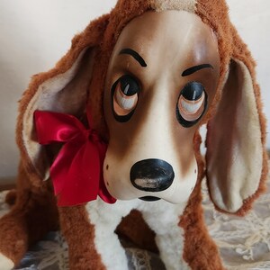 May include: A vintage plush dog toy with a brown and white body, large floppy ears, and a red satin bow. The dog has a painted face with large, expressive eyes and a black nose. The toy is sitting on a lace surface.