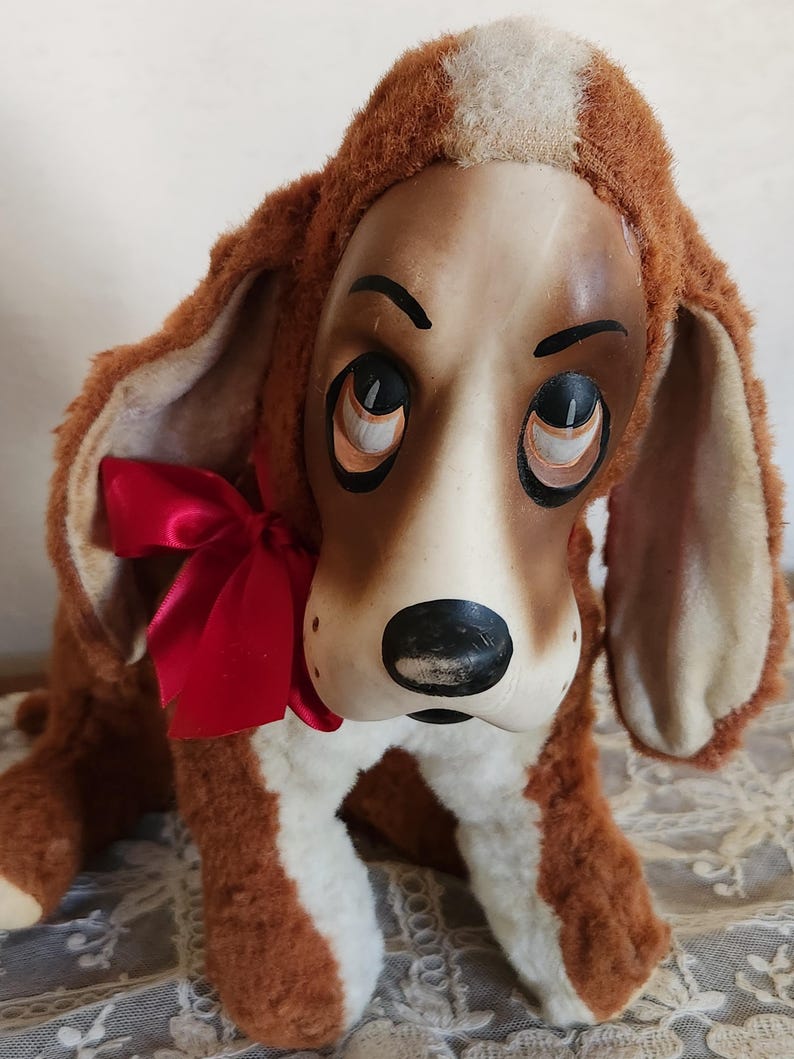 May include: A vintage plush dog toy with a brown and white body, large floppy ears, and a red satin bow. The dog has a painted face with large, expressive eyes and a black nose. The toy is sitting on a lace surface.