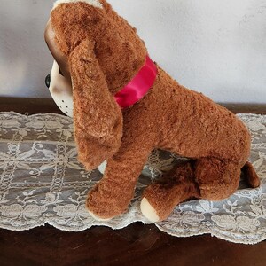 May include: A plush brown dog toy with floppy ears and a pink ribbon collar. The dog has a white patch on its head and white paws. The toy is sitting on a white lace tablecloth.