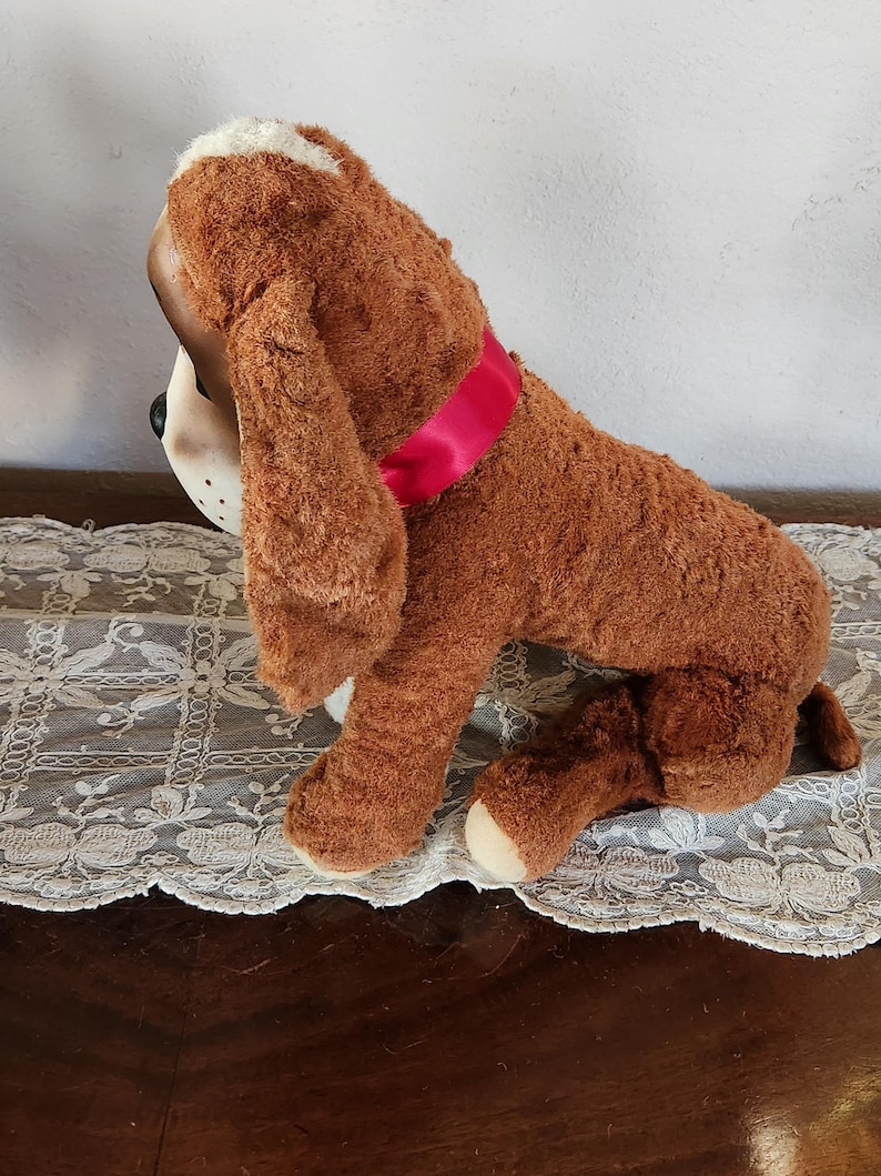 May include: A plush brown dog toy with floppy ears and a pink ribbon collar. The dog has a white patch on its head and white paws. The toy is sitting on a white lace tablecloth.