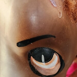 May include: Close-up of a doll's face, focusing on the eye and eyebrow. The eye is black with white and brown accents. The eyebrow is a thick, curved black line. The skin is a light brown color, and the doll has reddish-brown hair.