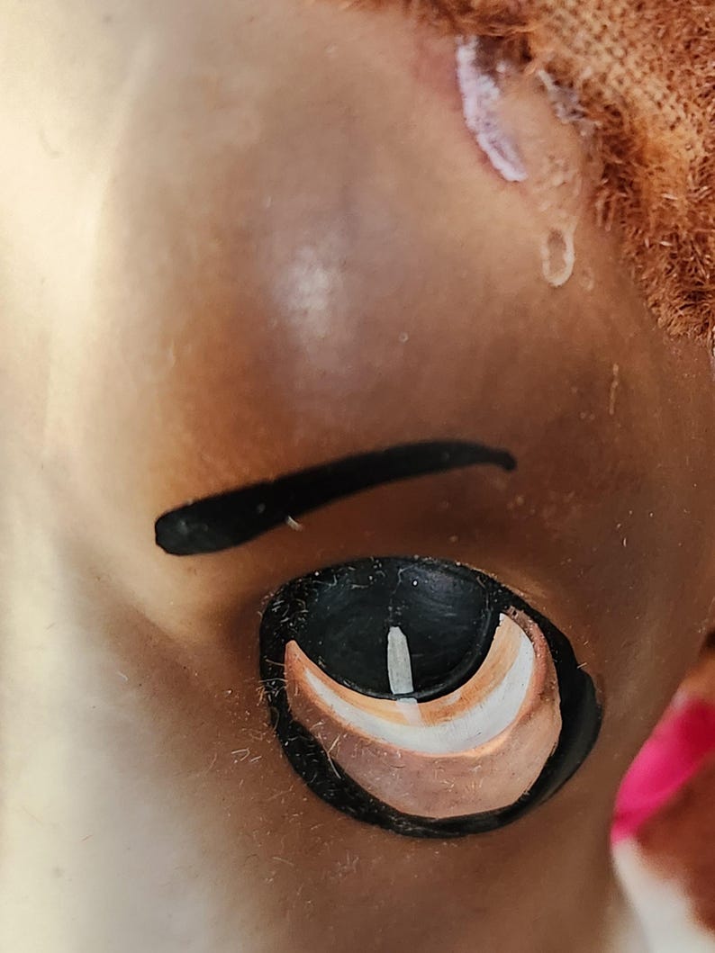 May include: Close-up of a doll's face, focusing on the eye and eyebrow. The eye is black with white and brown accents. The eyebrow is a thick, curved black line. The skin is a light brown color, and the doll has reddish-brown hair.