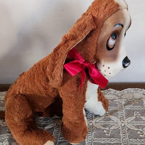 May include: A plush brown and white dog toy with long floppy ears and a red ribbon tied around its neck. The dog has a painted face with large eyes and a black nose. The toy is sitting on a decorative lace tablecloth.