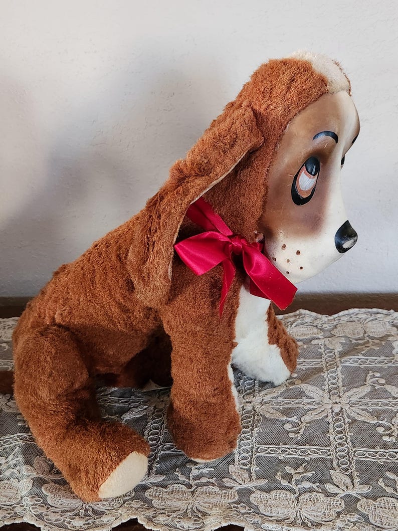 May include: A plush brown and white dog toy with long floppy ears and a red ribbon tied around its neck. The dog has a painted face with large eyes and a black nose. The toy is sitting on a decorative lace tablecloth.
