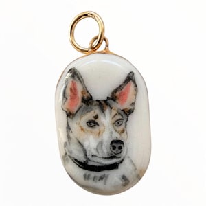May include: An oval-shaped pendant featuring a hand-painted portrait of a dog with white, brown, and black fur, pointed ears with pink inner details, and a black collar. The pendant has a smooth, glossy finish and a gold-toned metal jump ring.