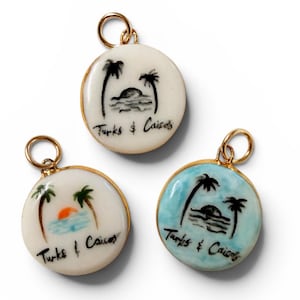 May include: Three round gold-rimmed pendants featuring hand-painted tropical scenes of palm trees, water, and the text "Turks & Caicos". One pendant has a white background, one has a light blue background, and one features a small orange sun.