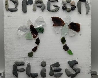 May include: A white wooden plaque with the words "DRAGON FLIES" spelled out using gray stones. Two dragonflies are depicted with sea glass wings in shades of brown, green, and clear. The plaque is propped up on a small wooden easel.