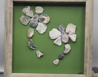 May include: A framed artwork featuring two floral arrangements made of various seashells on a green background. The seashells are in shades of white, gray, and tan. The frame is a light gray color. The artwork is displayed on a small wooden easel.