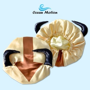 Puede incluir: Dos gorros de satén amarillo claro, con cuernos negros y un diseño de flecha marrón. Los gorros están diseñados para parecerse a un personaje de dibujos animados. El logotipo de Ocean Motion es visible en la esquina superior izquierda.