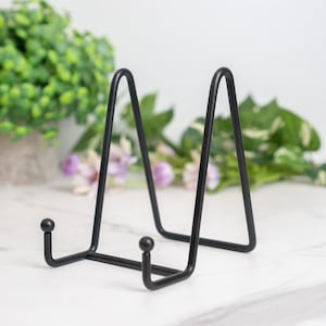 May include: A black metal display stand with a minimalist design. The stand has a triangular shape with a curved top and small ball feet. It is designed to hold plates or other decorative items. The stand is approximately 7 inches tall.