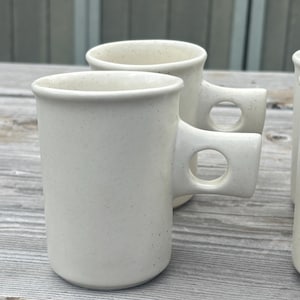 May include: A set of off-white ceramic mugs with a speckled finish. Each mug features a cylindrical body and a unique handle design that includes a circular cutout in the center, providing a distinct geometric look to the drinkware set.