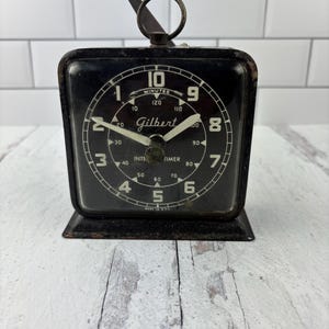 May include: Black vintage Gilbert Intertimer stopwatch with a white face and black hands. The stopwatch has a black metal case and a black metal base. The stopwatch is made in the U.S.A.