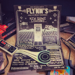 May include: A vintage poster advertising a new game at Flynn's, a retro arcade. The poster features a black and white pixelated image of a video game cabinet with the text "NEW GAME...LOADING..." The poster also lists other games available at the arcade, including "The End", "Intrude", "Zero Hour", "Matrix Blaster", "Code Wars", and "Derezzed".