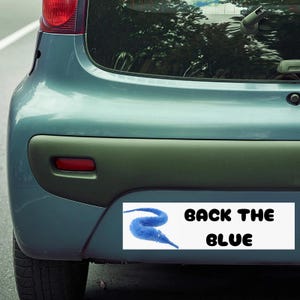 以下が含まれることがあります： 「BACK THE BLUE」の黒い文字が書かれた白い長方形のステッカーが付いた、水色の車のバンパー。ステッカーには青いミミズのようなデザインが施されています。バンパーは濃い緑色です。