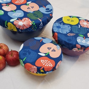 May include: Three blue fabric bowl covers with a colorful apple print design. The covers are on bowls of different sizes. The bowls are on a white surface.