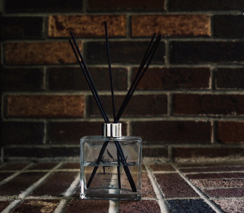 May include: A clear glass diffuser bottle with black reeds and a label that reads "gather round" and "our candle". The bottle is sitting on a brick surface.