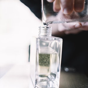 May include: A clear glass bottle with a silver cap is being filled with liquid from a glass pitcher. The bottle is sitting on a silver surface.