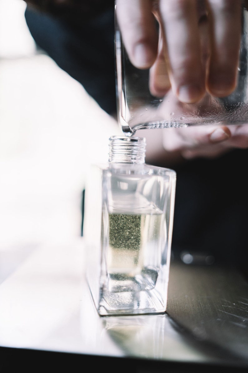 May include: A clear glass bottle with a silver cap is being filled with liquid from a glass pitcher. The bottle is sitting on a silver surface.