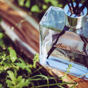 May include: A clear glass bottle with a silver cap and black diffuser sticks. The bottle is labeled "gather 'round come a'round".
