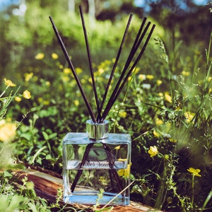 May include: A clear glass bottle with black diffuser sticks, filled with a clear liquid. The bottle is sitting on a wooden surface surrounded by green grass and yellow flowers.
