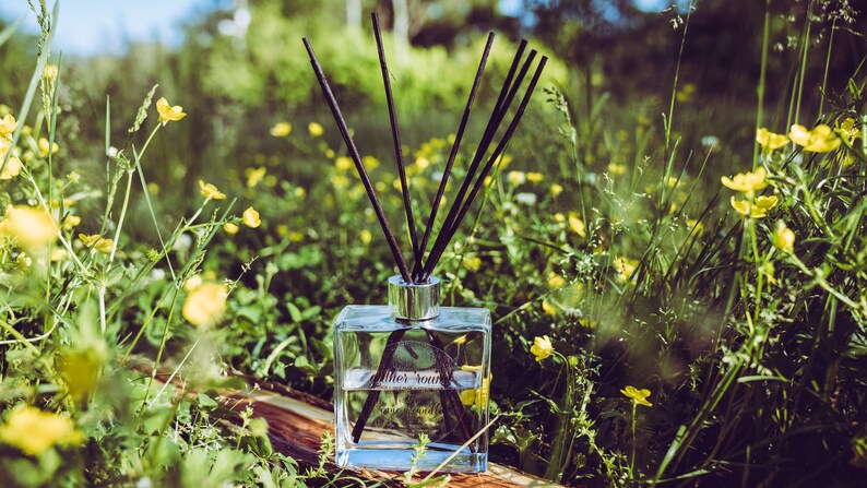 May include: A clear glass bottle with black diffuser sticks, filled with a clear liquid. The bottle is sitting on a wooden surface surrounded by green grass and yellow flowers.