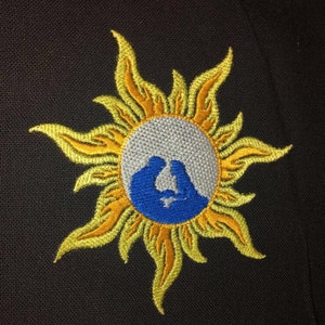 May include: Embroidered patch featuring a stylized sun with yellow and orange rays. The center circle has a gray background with a blue silhouette of two figures. The patch is on a black fabric background.