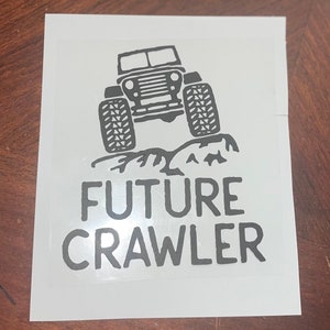 Peut inclure: Autocollant noir et blanc représentant un véhicule tout-terrain avec de gros pneus, grimpant sur un terrain rocheux. Le texte "FUTURE CRAWLER" est imprimé en dessous du véhicule dans une police de caractères grasse et sans empattement. L'autocollant est sur fond blanc.