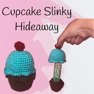 May include: Two crocheted cupcake toys. One is a complete cupcake with a brown base, turquoise frosting, and a red cherry on top. The other is a cupcake with a turquoise frosting top, a green slinky, and a brown base. The text "Cupcake Slinky Hideaway" is at the top.