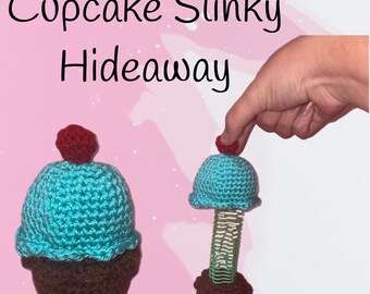 May include: Two crocheted cupcake toys. One is a complete cupcake with a brown base, turquoise frosting, and a red cherry on top. The other is a cupcake with a turquoise frosting top, a green slinky, and a brown base. The text "Cupcake Slinky Hideaway" is at the top.