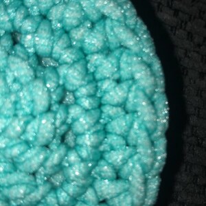 May include: Close-up of a round, turquoise crochet item. The texture is thick and the stitches are visible. The item has a slightly shiny appearance, with highlights catching the light. The background is dark and out of focus.