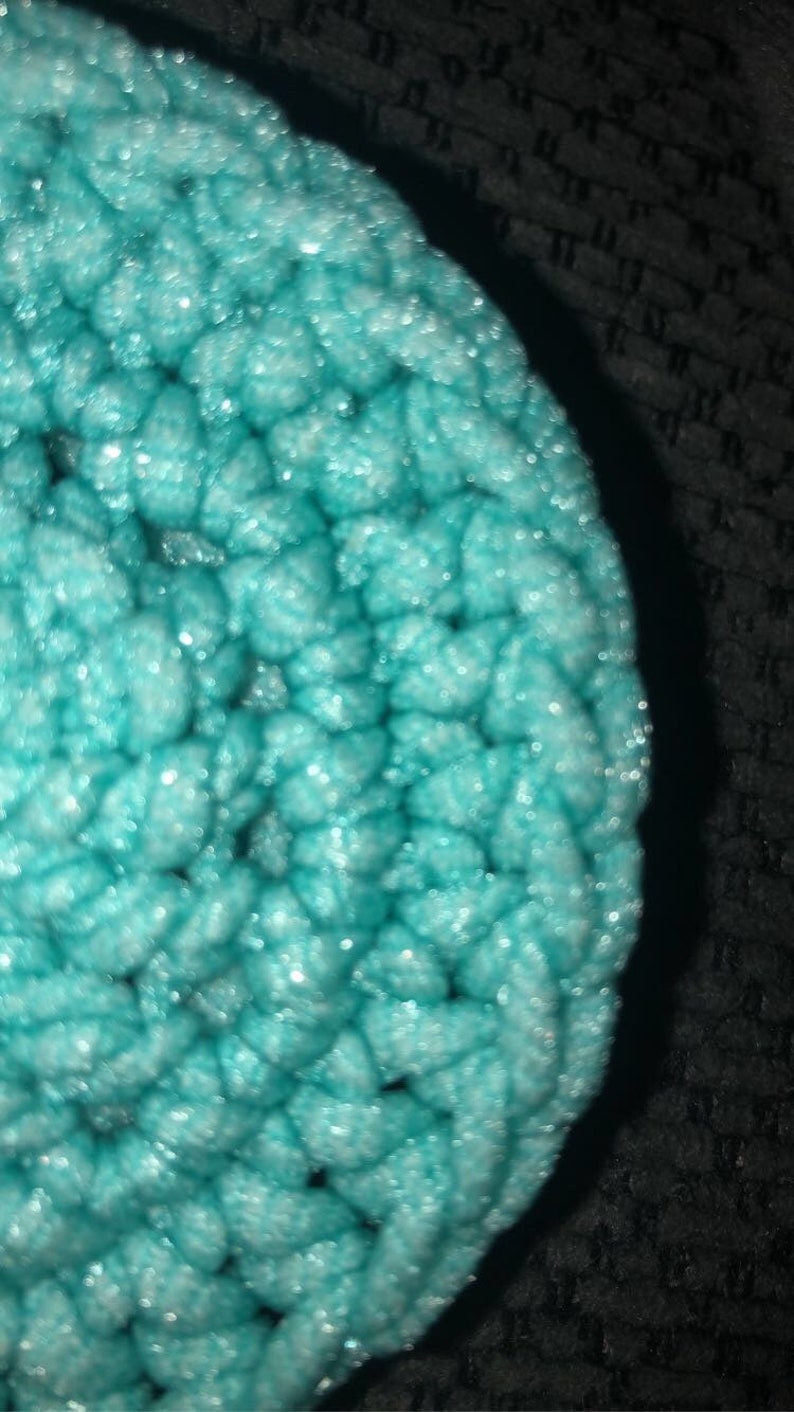 May include: Close-up of a round, turquoise crochet item. The texture is thick and the stitches are visible. The item has a slightly shiny appearance, with highlights catching the light. The background is dark and out of focus.