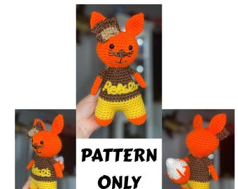 May include: Crocheted fox toy in orange, brown, and yellow, with the text "Reese's Cup Fox" above the image. The fox has a bow, a brown shirt with "Reese's" on it, and yellow shorts. The text "PATTERN ONLY" is also visible.